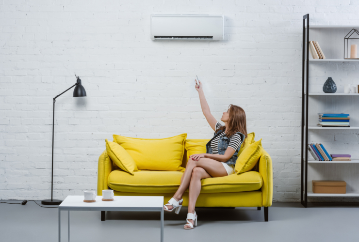 attractive young woman sitting on sofa and pointing at air conditioner with remote control